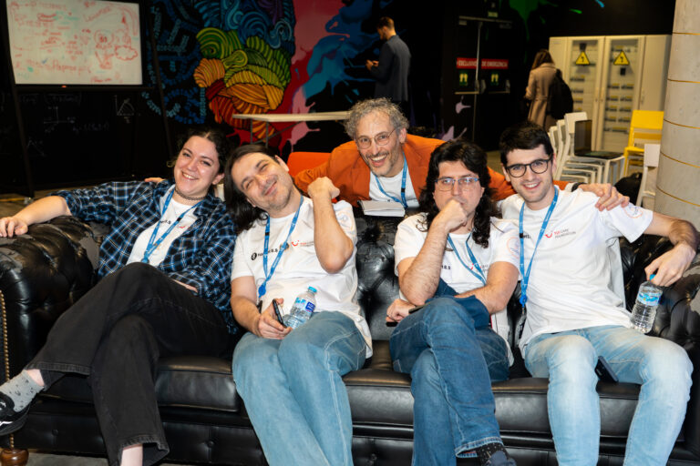 Four people sitting on a sofa and smiling, another one standing behind the sofa leaning down towards the others for a group picture