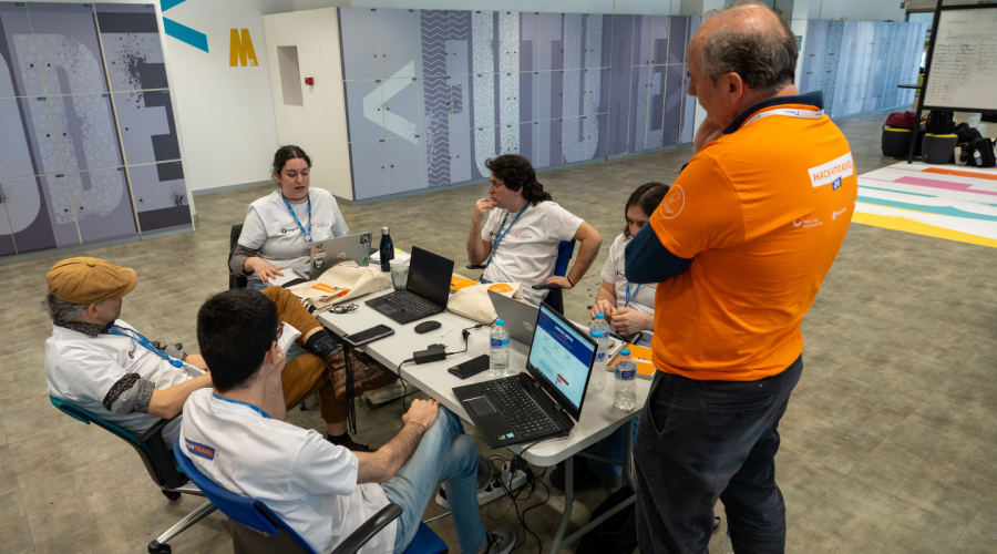 a group of people sitting on a table in front of their computers talking to a men standing next to them